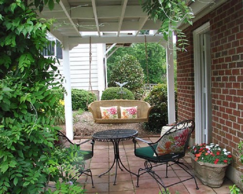 A cozy patio with a swing, two chairs, and a small table, surrounded by greenery and flowers.