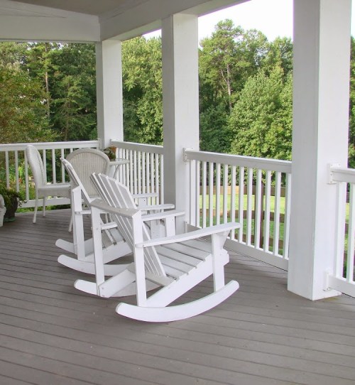 Two white rocking chairs on a porch overlooking a green landscape, surrounded by trees.