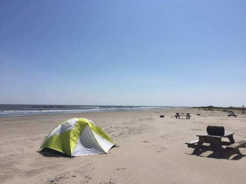 A bright beach scene featuring a green and white tent, picnic tables, and calm waves under a clear blue sky.