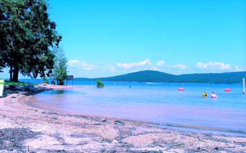 A serene lakeside scene with clear blue water, sandy shore, and distant hills under a bright sky. Two people are swimming.