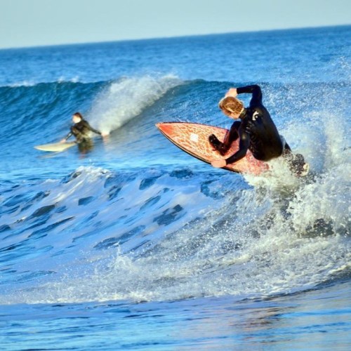 Two surfers riding waves in the ocean, one performing a maneuver on a red surfboard. Bright blue water in the background.
