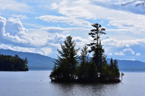 A serene lake scene featuring a small island with trees, surrounded by calm water and a backdrop of clouds and mountains.