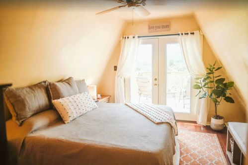 Cozy bedroom with a large bed, light curtains, and a potted plant, featuring a balcony door and warm yellow walls.