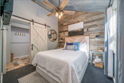Cozy bedroom with a wooden accent wall, a queen bed, and rustic decor, featuring a ceiling fan and sliding barn door.