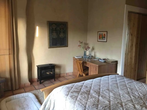 Cozy bedroom with a wooden bed, desk, and a small fireplace, featuring warm lighting and simple decor.
