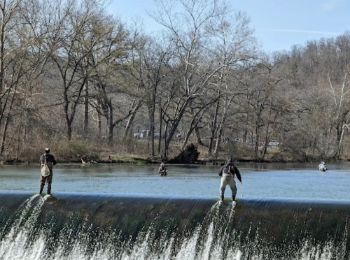 Two fishermen stand on a waterfall edge, while others fish in the river surrounded by bare trees and a clear sky.