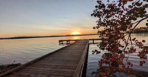 A serene sunset over a calm lake, with a wooden dock and autumn leaves in the foreground.