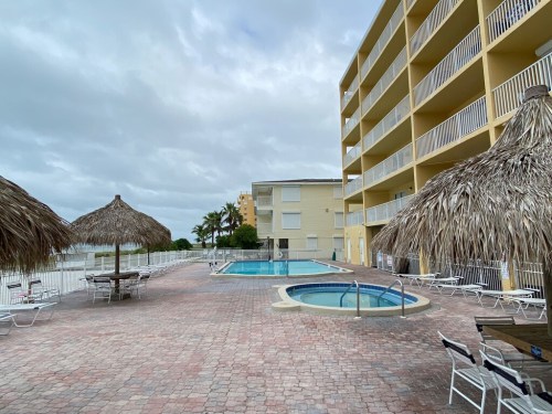 A pool area with a hot tub, surrounded by palm trees and thatched umbrellas, next to a yellow building under cloudy skies.