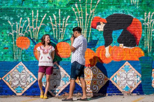 A woman and man stand in front of a colorful mural, smiling and interacting in a vibrant outdoor setting.