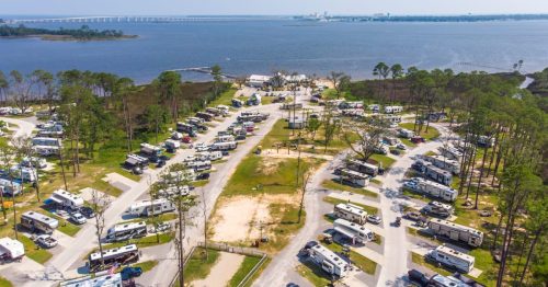 Aerial view of a large RV park with numerous parked RVs, surrounded by trees and a body of water in the background.