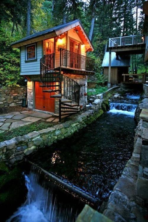 A charming two-story cabin with a spiral staircase, surrounded by trees and a flowing stream with small waterfalls.