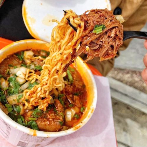 A close-up of a fork lifting noodles from a bowl of spicy soup with meat and herbs.