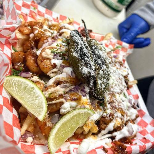 A colorful platter of loaded fries topped with shrimp, jalapeños, lime, and drizzled with sauces on a checkered paper.