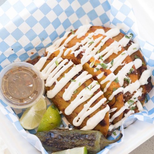 A basket of crispy tacos drizzled with sauce, served with lime, a jalapeño, and a small container of salsa.