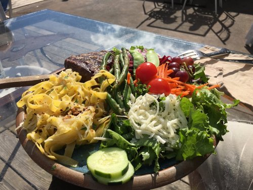 A colorful plate featuring pasta, grilled steak, mixed greens, cucumbers, tomatoes, and assorted vegetables.