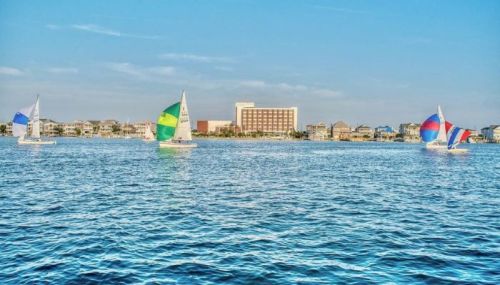 Three sailboats with colorful sails glide across a calm blue sea, with a hotel and shoreline in the background.