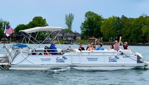A group of people enjoying a boat ride on a lake, waving and smiling, with trees and houses in the background.
