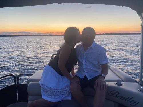 A couple sharing a kiss on a boat at sunset, with water and a colorful sky in the background.
