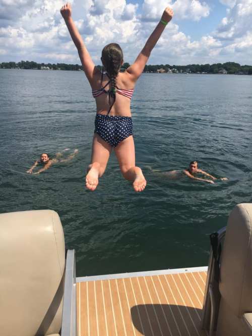 A girl jumps off a boat into the water, while two others swim nearby on a sunny day.