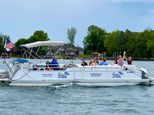 A group of people enjoying a boat ride on a lake, waving and smiling, with trees and houses in the background.
