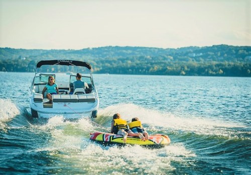 A boat pulls two people on a colorful inflatable tube across a lake, with a scenic backdrop of hills and blue sky.