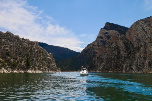 A boat glides through calm waters surrounded by steep, rocky cliffs under a blue sky with scattered clouds.