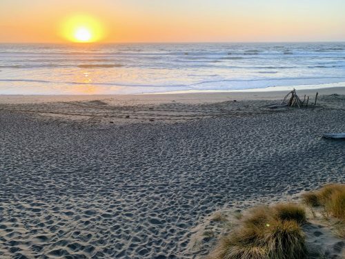 Sunset over a calm ocean, casting warm hues on the sandy beach with gentle waves and sparse vegetation in the foreground.