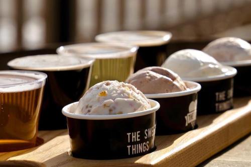 A wooden tray holds cups of ice cream and glasses of beer, showcasing a variety of flavors and colors in sunlight.