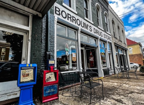 Exterior of Borroum's Drug Store, featuring a vintage storefront, newspaper stands, and outdoor seating.