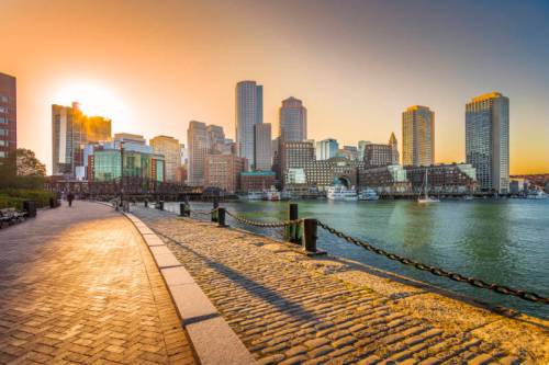 Sunset over a city skyline with tall buildings, reflecting on water, along a cobblestone waterfront path.