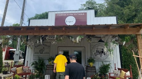 Two people walk towards a rustic farm-to-table restaurant with a sign reading "Famoda Farm to Table." Overcast sky.