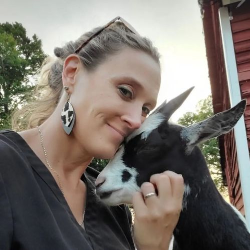 A woman smiles while gently petting a goat, with a rustic background of trees and a red barn.