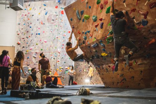 Two climbers bouldering on a wall in a climbing gym, with spectators sitting nearby and colorful holds on the wall.