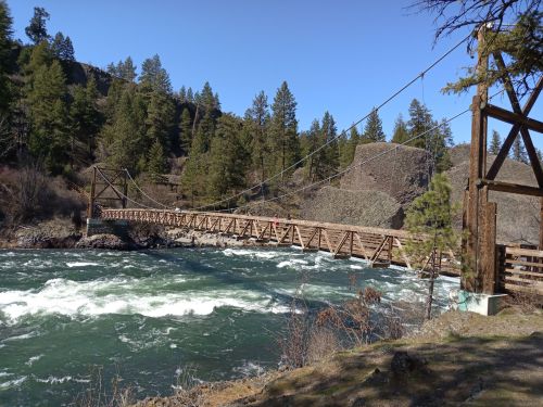 A wooden suspension bridge spans a rushing river, surrounded by lush green trees and rocky terrain under a clear blue sky.