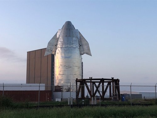 A shiny rocket stands tall against a clear sky, with a large building in the background and a fenced area in the foreground.