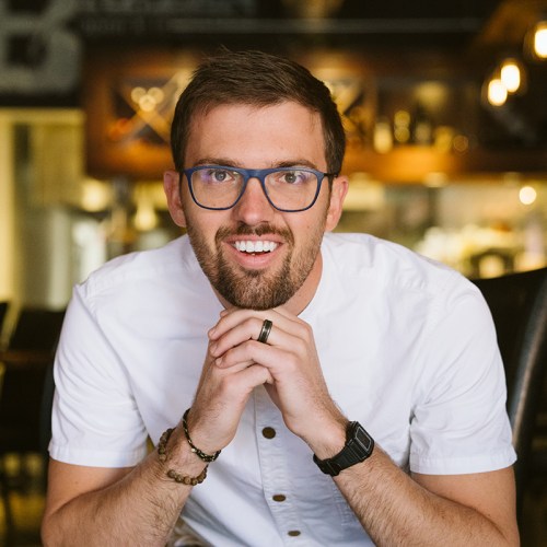 A smiling man with glasses and a white shirt sits at a table, resting his chin on his hands in a cozy setting.