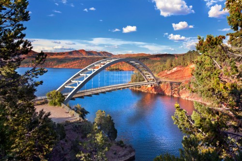 A modern arch bridge spans a serene lake, surrounded by trees and mountains under a blue sky with fluffy clouds.