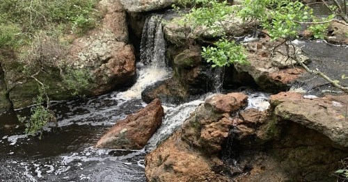 A serene waterfall cascading over rocks into a dark pool, surrounded by lush greenery.