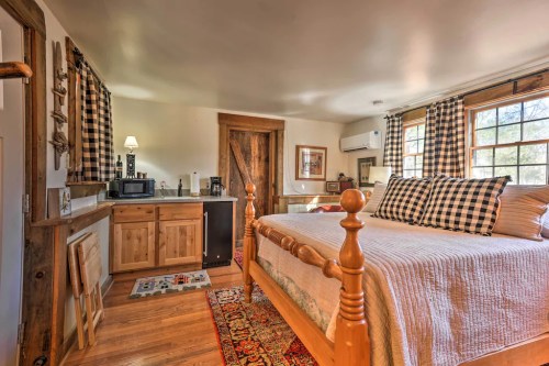 Cozy bedroom with a wooden bed, kitchenette, and large windows, featuring rustic decor and checkered curtains.