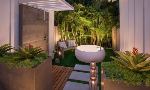 A serene outdoor bathroom with a round tub, shower, and lush greenery, surrounded by decorative plants and soft lighting.