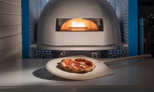 A freshly baked pizza on a wooden peel in front of a wood-fired oven with flames visible inside.