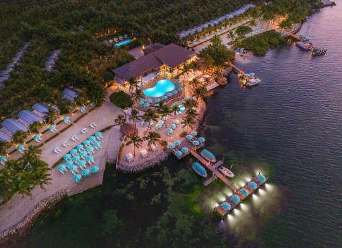 Aerial view of a tropical resort with a pool, cabanas, and boats along a serene waterfront at sunset.