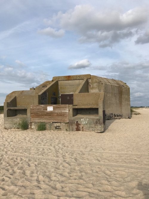 A weathered concrete bunker on a sandy beach, with a "Keep Off" sign and cloudy skies above.