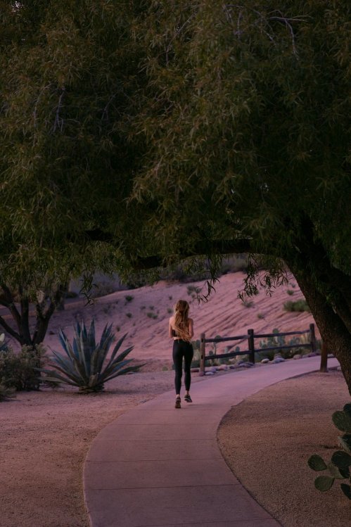 A woman walks along a winding path surrounded by trees and desert vegetation at dusk.