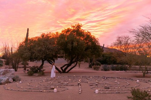 A person in a white dress stands in a stone labyrinth at sunset, surrounded by desert plants and a colorful sky.