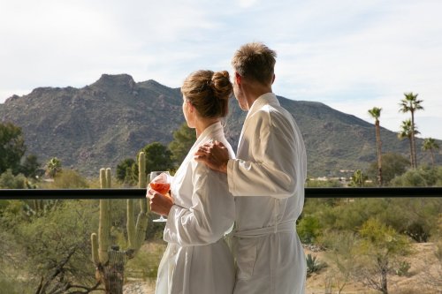 A couple in bathrobes stands on a balcony, gazing at a mountain landscape with palm trees and cacti.