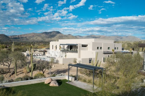 Modern white house surrounded by desert landscape, mountains in the background, and a clear blue sky.