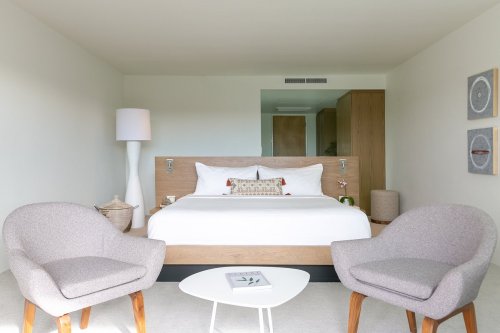 A modern hotel room featuring a bed, two chairs, a small table, and minimalist decor in neutral tones.
