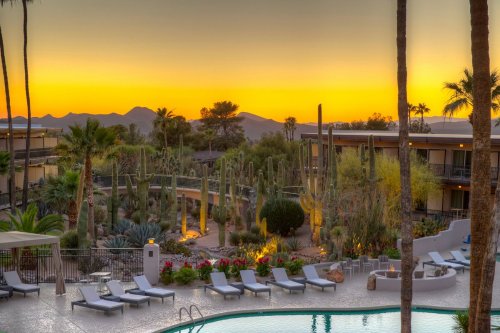 A serene resort scene at sunset, featuring a pool, lounge chairs, palm trees, and mountains in the background.