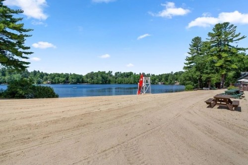 A sandy beach by a calm lake, with a lifeguard tower and trees lining the shore under a clear blue sky.